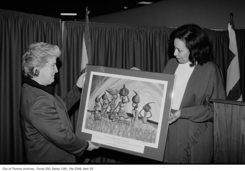 A woman of African descent and a white woman are together holding a framed painting of African women in striped dresses carrying pots on their heads.