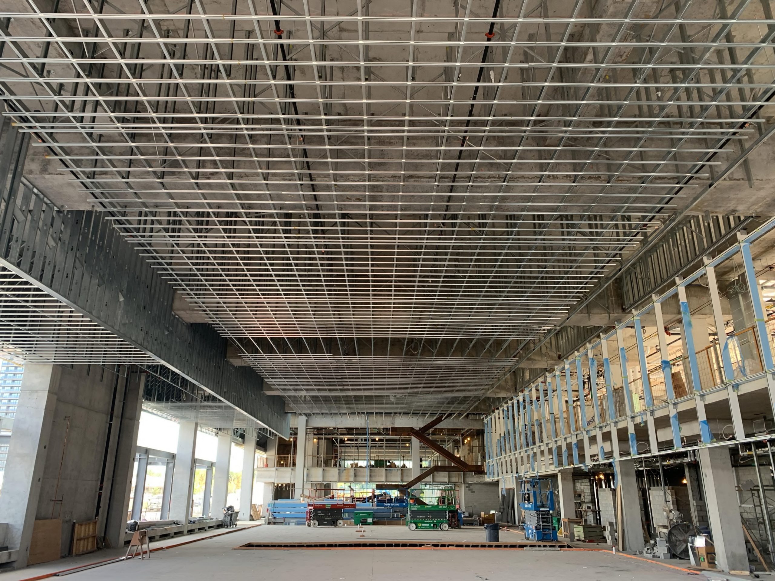 A picture of the interior of the new Community Recreation Centre while under construction. The pictures shows large exposed concrete walls with metal beams forming a grid across the ceiling. The second level is on the right side and does not have walls/glass windows yet. Various construction equipment is seen throughout the space. 