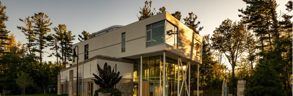 Colour photograph of the Clark Centre for the Arts taken at dusk, with trees in the background, and the evening sun highlighting the right side of the building creating golden highlights.