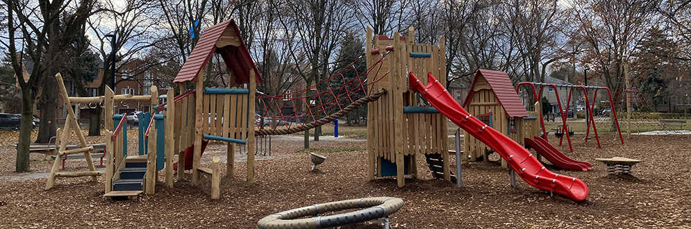 An image of Florence Gell Park Playground taken at ground level which shows the wooden junior and senior play structures (shown in red) in the background. The play area is surrounded by tall mature trees and grass.