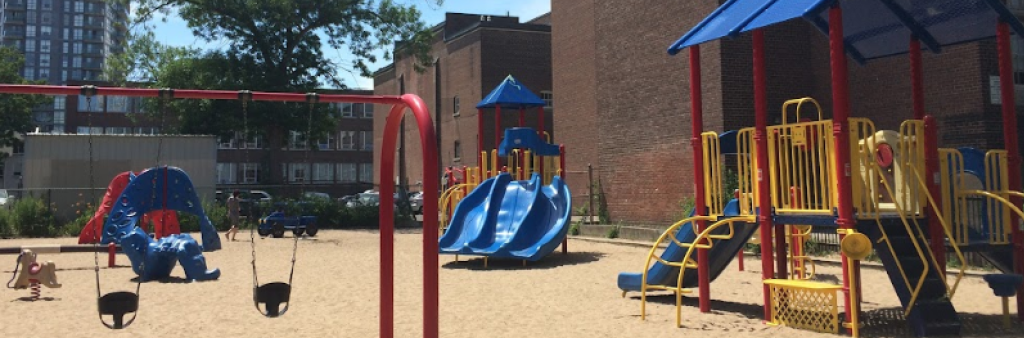 An image of the playground taken in 2018 which shows a swing set in the foreground and two blue, yellow and red play structures in the background. The playground is on top of sand and is right beside the University of Toronto Schools (UTS) building.