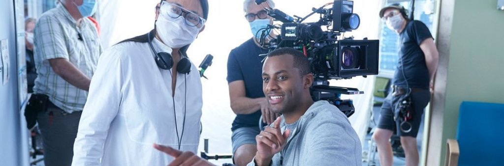 Actor pointing at something while talking to a film crew in a medical mask with camera operator in the background.