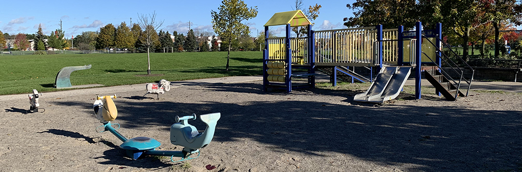 A photograph of the play equipment in Bill Hancox Park Playground, which includes a senior play structure with double slide and play panels and various stand-alone spring toys on top of sand. A large open lawn space surrounds the playground.