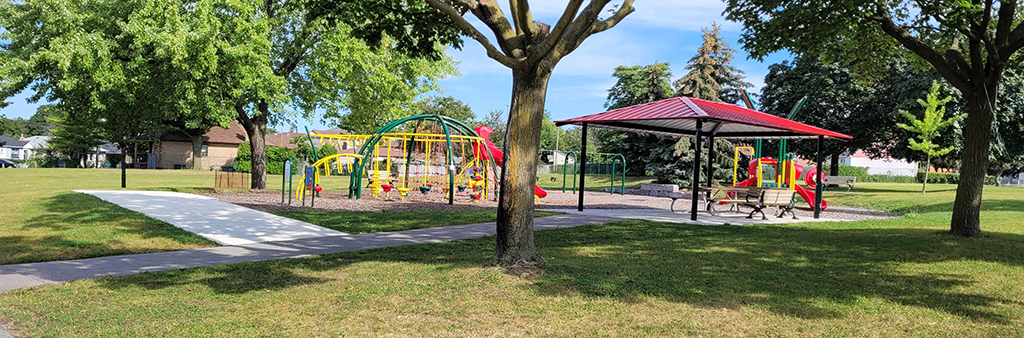 An image of the playground in Maple Leaf Park on a sunny day. The playground is surrounded by mature trees and open lawn space. The new splash pad will be located near the playground.