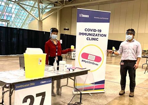 Two people at a City of Toronto immunization clinic, wearing face masks and face shields while pointing at the City's COVID-19 Immunization Clinic banner