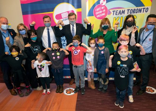 Mayor Tory with group of adults and children flexing arms in front of Team Toronto Kids sign at a City vaccine clinic