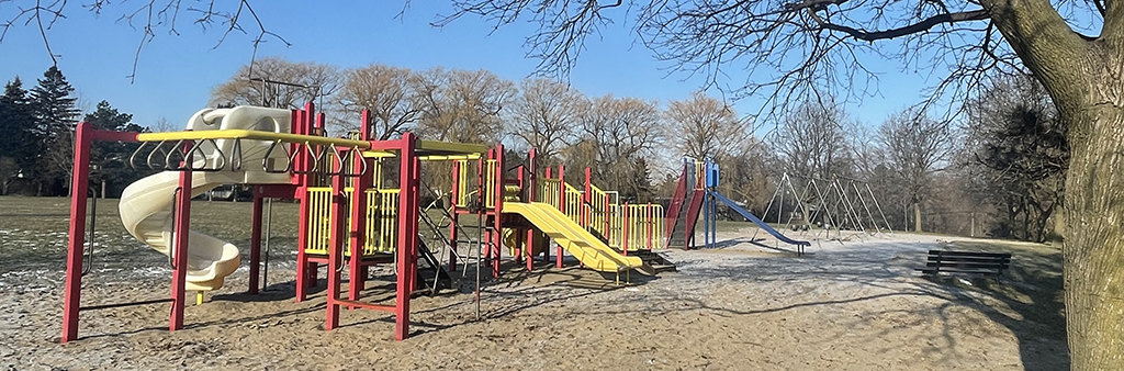 A photograph of Cedargrove Park Playground. The spiral slide and monkey bars are in the foreground, with a swing set in the background. The play equipment is on top of sand and the playground is surrounded by mature trees and open lawn.