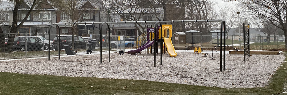 A photograph of S.A.D.R.A Park with a combined junior and senior play structure in the foreground and a swing set and climbing structure in the background. The play features are on top of sand.