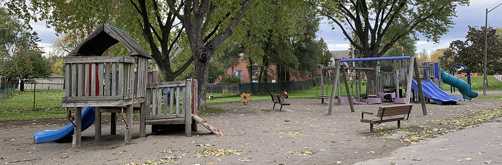 A photo of Benner Park's playground. It includes a climbing structure and swings, both made from wood. The area is surrounded by mature trees.