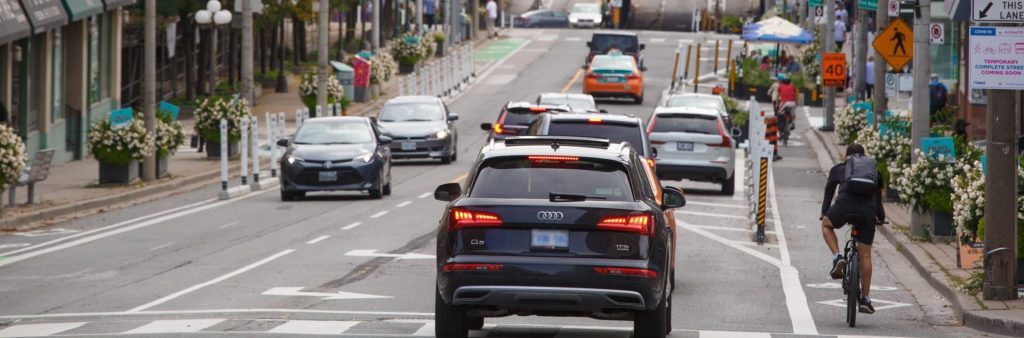 People and cycling on Yonge Street south of Woodlawn Avenue with two lanes of motor vehicle traffic and cycle tracks. The cycle tracks moves around a curbside patio set up as part of CafeTO. Planter line the sidewalks.