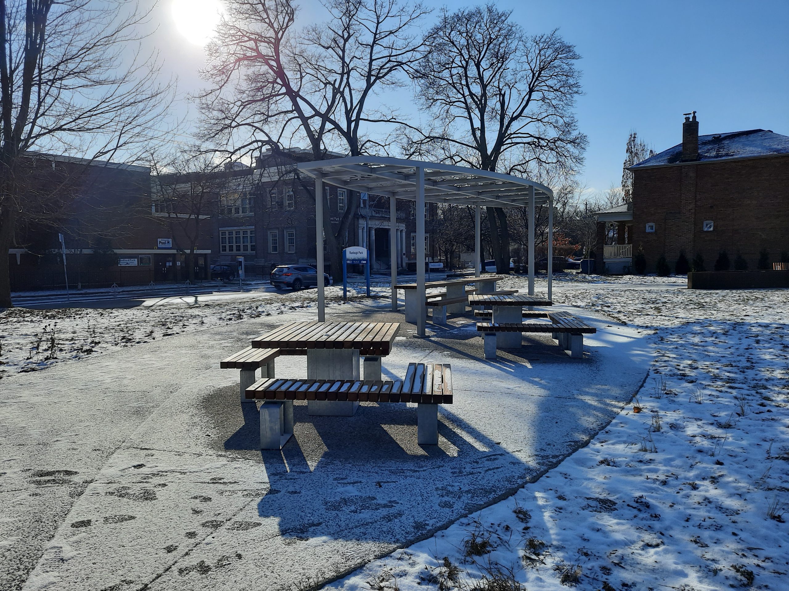 Photograph of the new Ranleigh Park showing new seating area with custom shade structure, picnic table, and games tables, with mature trees in the background.