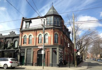 A corner building designed in the High Victorian style that features highly-patterned elevations using red and buff brick, distinctive semi-circular-headed windows, and a spire