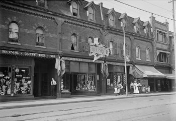 People walk on the sidewalk in front of storefront display windows that are filled with goods in a black and white photo from 1919