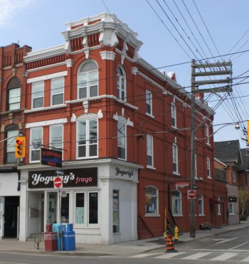 A red brick building with arched windows and white stone details sits prominently at the intersection of Queen Street West and Palmerston Avenue