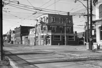 Illuminated by afternoon sunshine, a two-and-a-half storey building with a mansard roof stands next to a row of historic buildings across the empty intersection of Queen Street West and Ossington Avenue