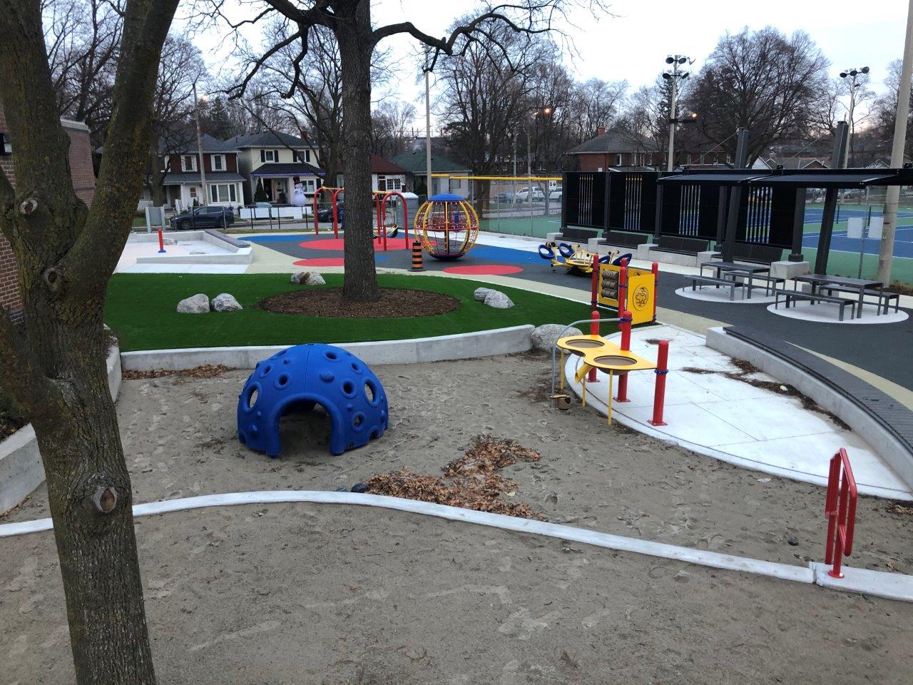 New playground at Trace Manes park. Image shows a view of park looking east and shows the sand play area with Cozy Dome, artificial turf area, with swings and spinning climber in the distance.
