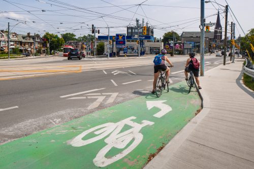 Two people on bikes wait in the green left turn bike box. The box contains a painted bicycle symbol and arrow pointing left.