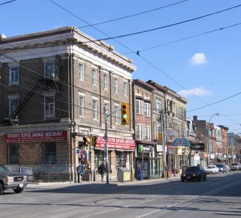 An old building with ornate stone details at its roof line sits prominently at the intersection of Queen Street West and Brock Avenue