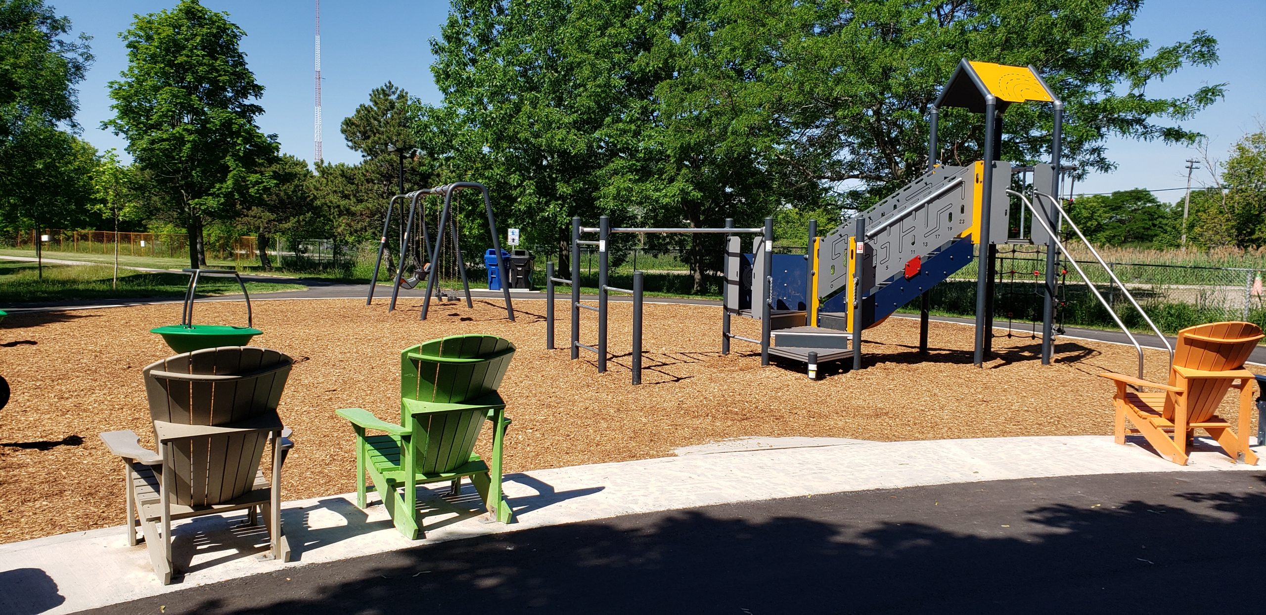 New play equipment at Natal Park, including climbing structure, accessible swing set and balance spinner on a wood fiber play surface. Play area is bordered by a paved pathway and Muskoka chairs.