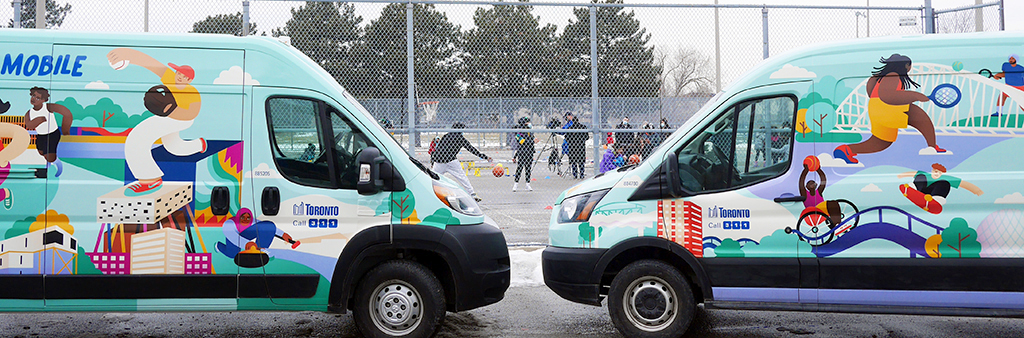 Two Play Mobile vans, covered in bright illustrations of people playing sports, are parked at Malvern Community Recreation Centre. In the background, people are playing basketball and other sports.