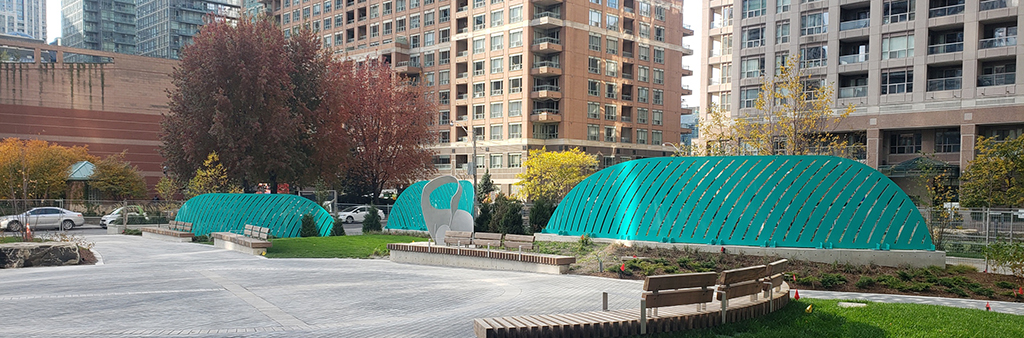 Image of the new Dr. Lillian McGregor Park show a central circular gathering space bordered by wooden benches, lawn and planting areas. Three green 'reed' screens are staggered in the background, as well as one white crane sculpture.