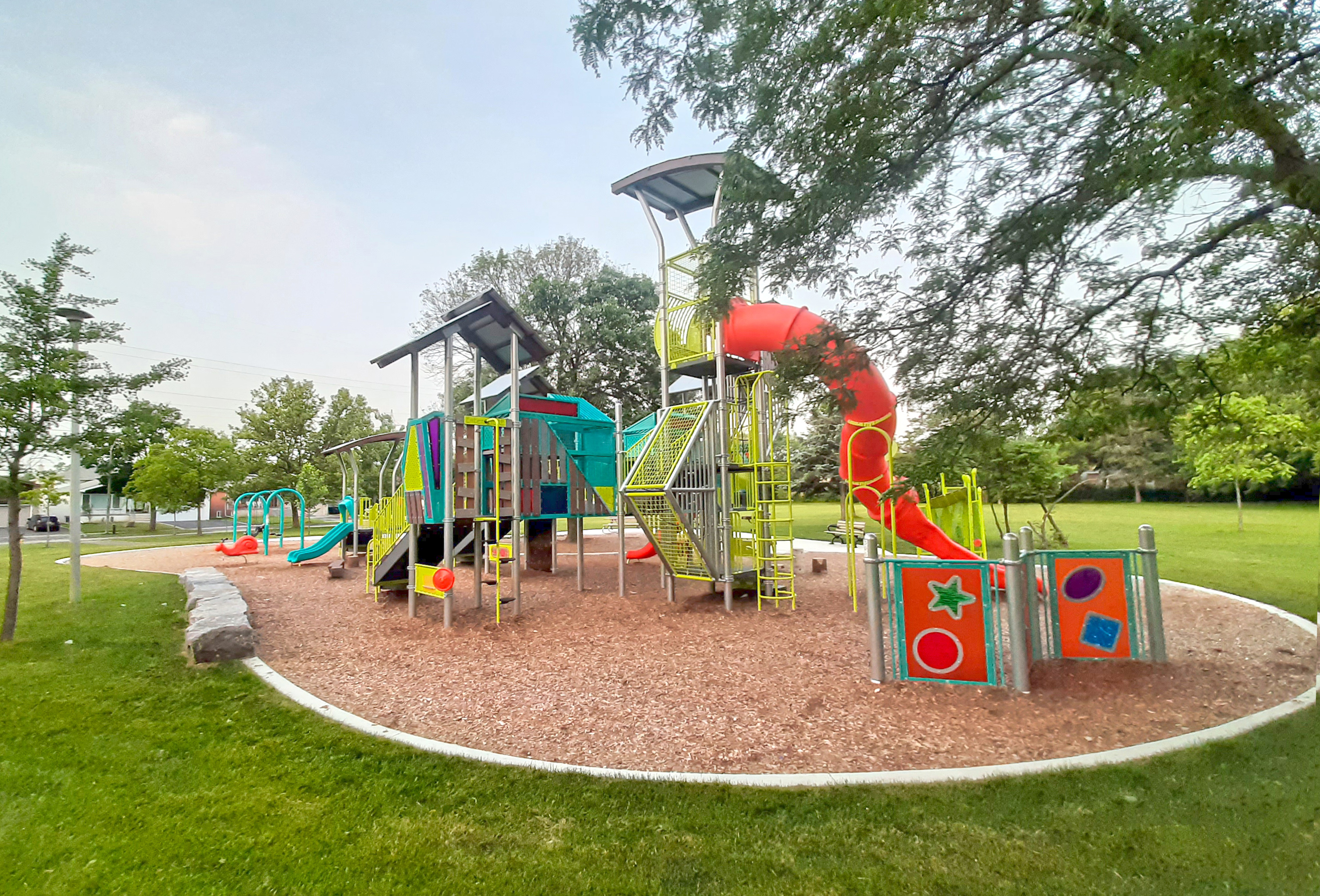 New play equipment at Charlton Park features brightly coloured climbing structures and slides of various heights. Swing set and other play features are also visible. All play equipment sits on a wood fiber play surface.