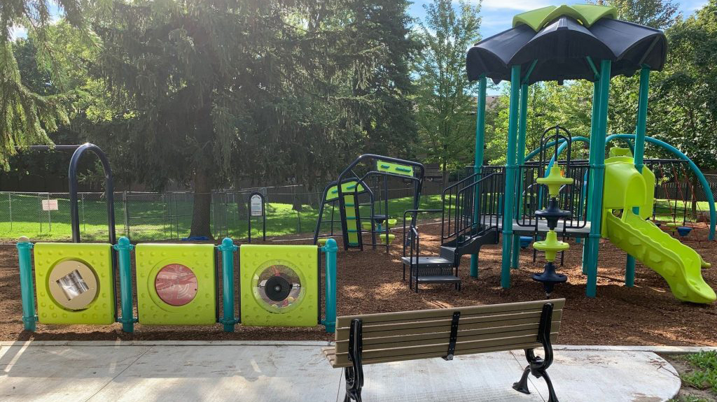 New play equipment at Old Sheppard Park features brightly coloured climbing structures and activity panels on a wood fiber play surface. A paved area with a bench faces the play area.
