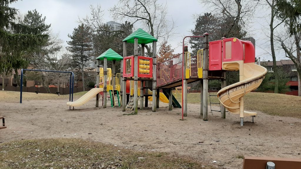 Old wood and plastic playground equipment, including three yellow slides of various sizes, and a swing set on a sandy play surface.