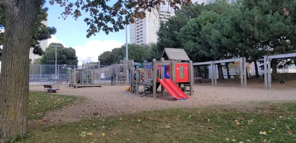 Old playground equipment at Roywood Park includes wooden junior and senior play structures with plastic slides and three wooden swing sets on a sandy play surface.