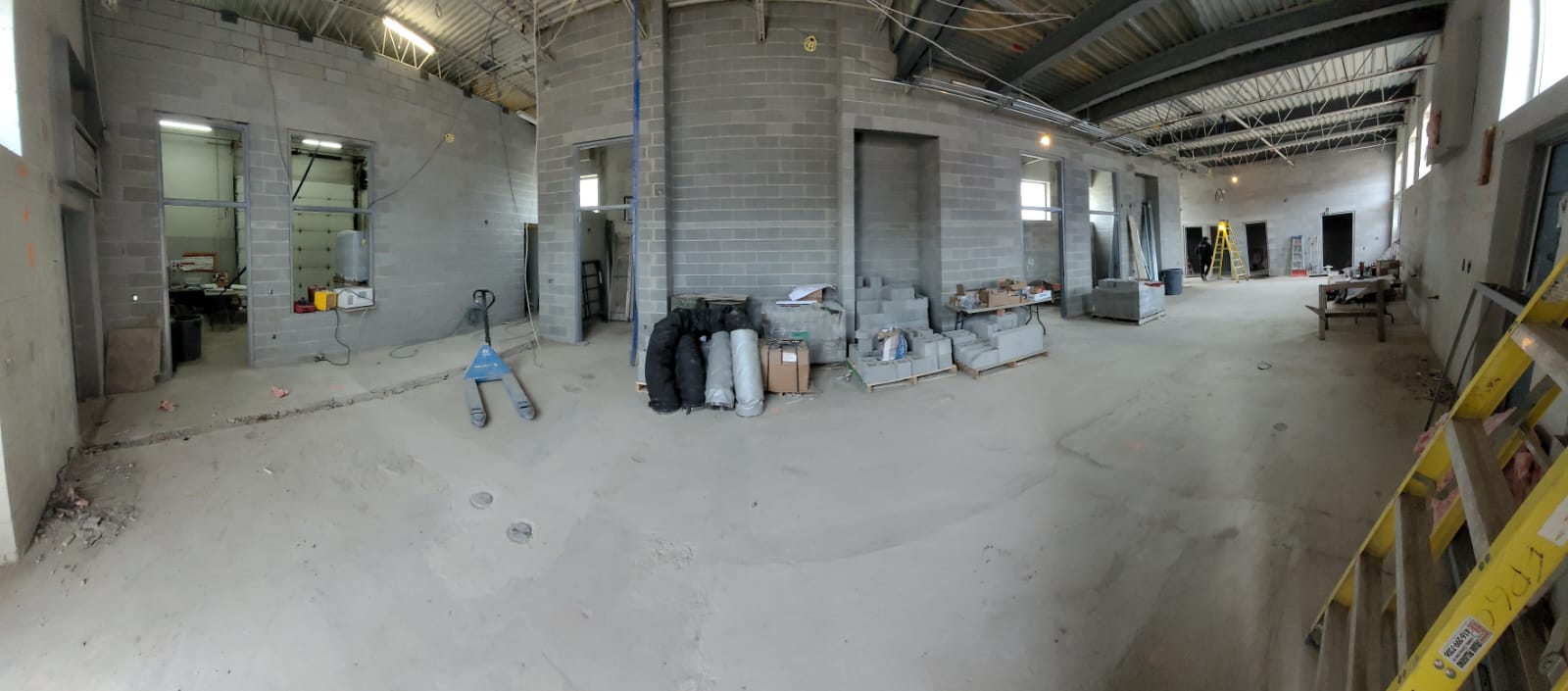 Grey cinderblock walls and door frames define an office space and other small rooms from the main clubhouse space. Ladders and other equipment and materials are scattered throughout the room on an unfinished cement floor.