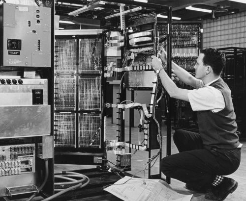 Black and white view of room with four large computer panels. Man crouching down adjusting wires on one of the panels.