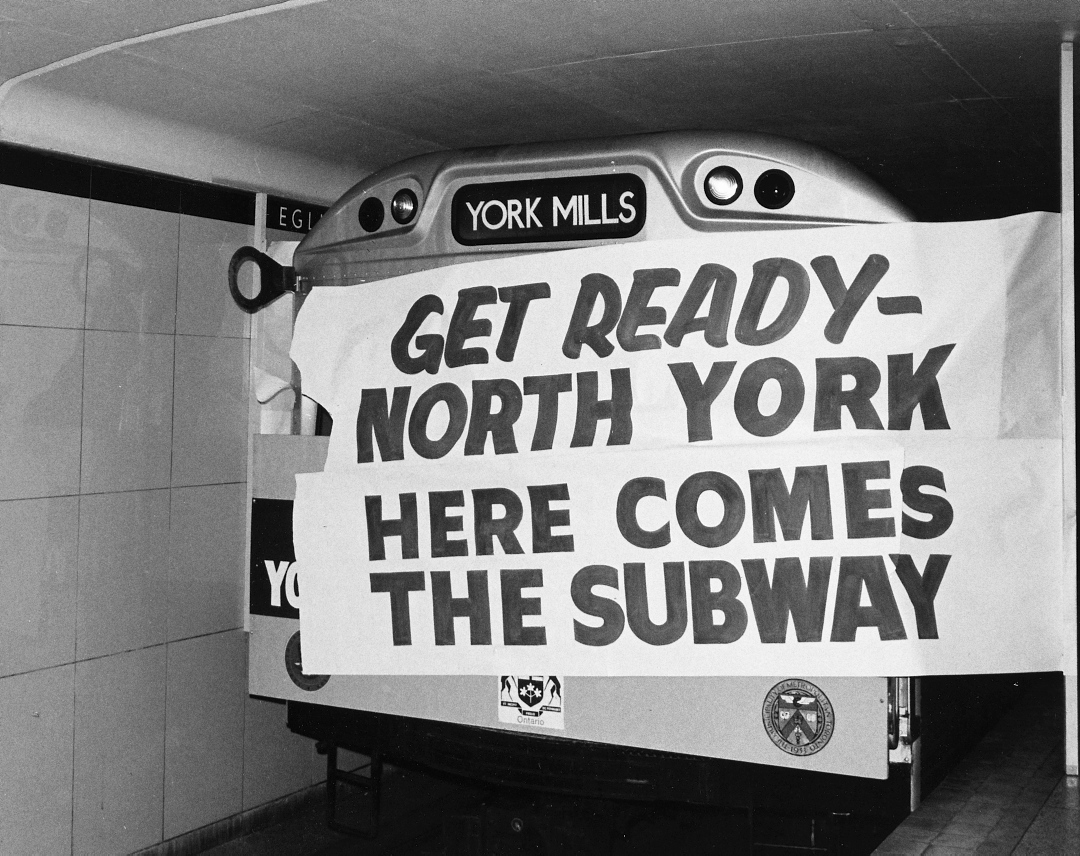 Black and white view of subway train breaking through a painted paper sign that reads Get Ready North York Here Comes the Subway