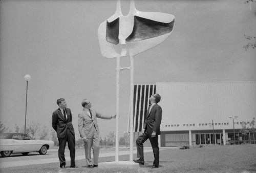 Black and white photo of three men looking at a large outdoor sculpture in front of a large white building. 