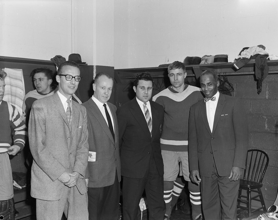 Black and white view of seven men in a locker room, four wearing suits and three in hockey uniforms.