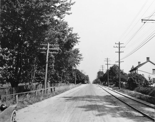 View of dirt road with rail line, hydro line and a house. 