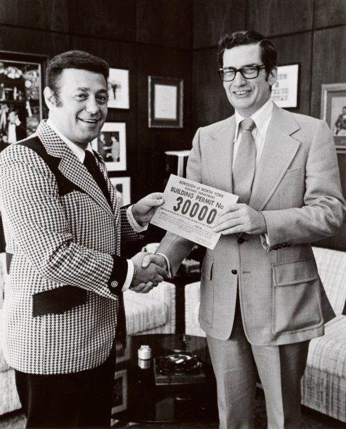 Two men in suits shaking hands and jointly holding a piece of paper which reads Building Permit No. 30000