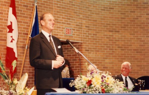 Colour view of man in suit standing in front of microphone with flags and brick wall behind him.