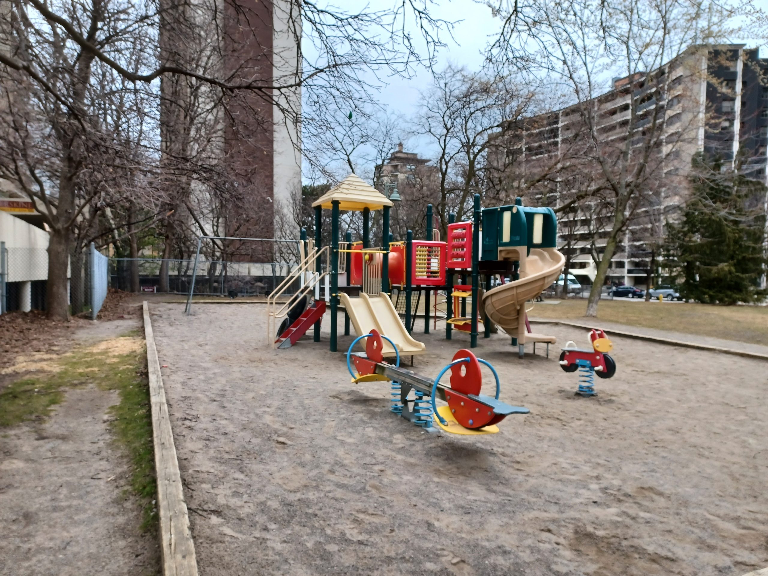 Redpath Avenue Parkette playground, which shows a teeter-totter and spring toy in the foreground and medium-sized play structure and swing set in the background. The playground is on top of sand and includes a short wood border around the entire playground.