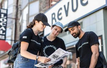 Three people wearing Myseum t-shirts looking at a newspaper in front of the Revue theatre