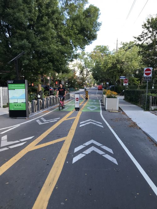 A person cycles through a cycling-only block that features directional arrows in opposite lanes, planters, a Bike Share station, and "bicycles excepted" sign. 
