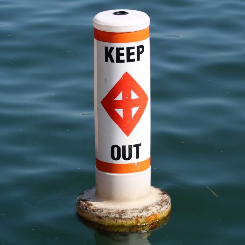This is a photo of a keep out buoy in the lake. The buoy is white with two horizontal orange bands and an orange cross inside an orange diamond on two opposite sides.