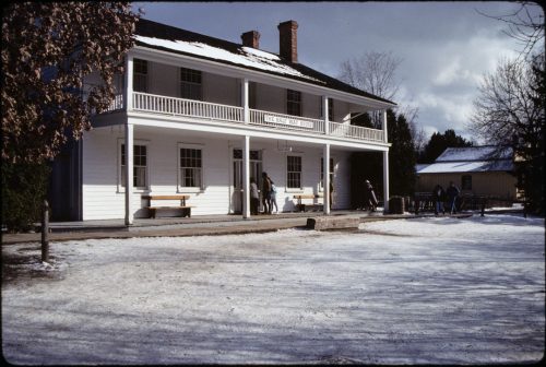 Colour view of white wooden two story building in winter with snow in foreground.