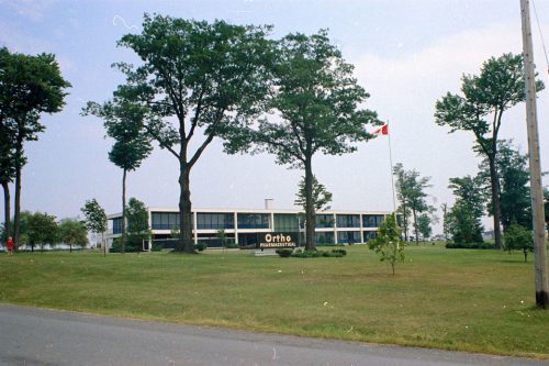 Colour exterior view of long, two story commercial building with Ortho Pharmaceutical sign, trees and grass in foreground.