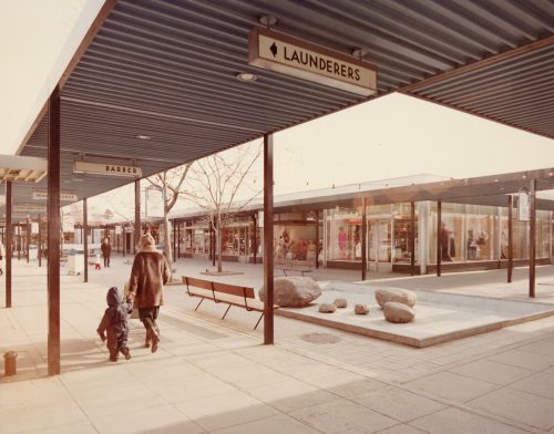 Colour view of outdoor shopping plaza with woman and child walking. 