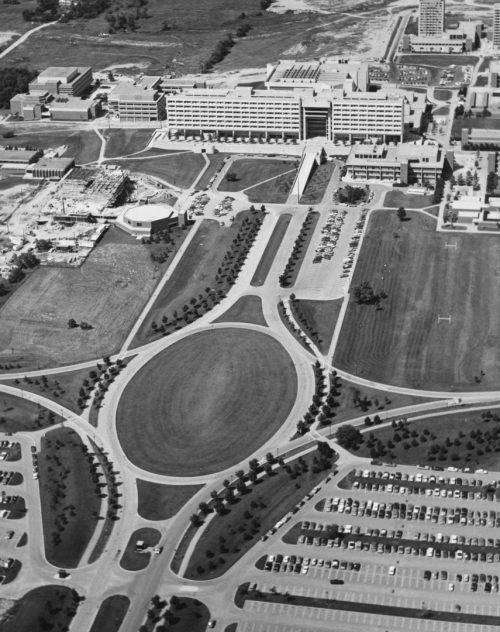 Black and white oblique aerial view of York University campus buildings, parking lot and large, oval landscaped green space.