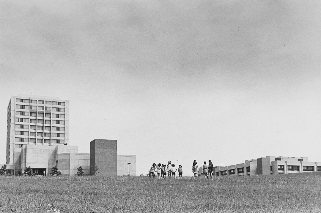 Black and white view of a group of about ten young people in grassy field with three university campus buildings in background.