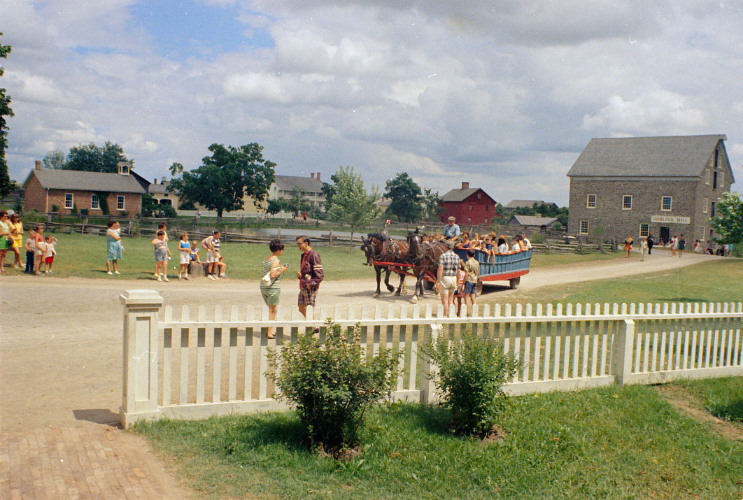 Colour view of historic buildings and people on horse-drawn wagon with fence in foreground.