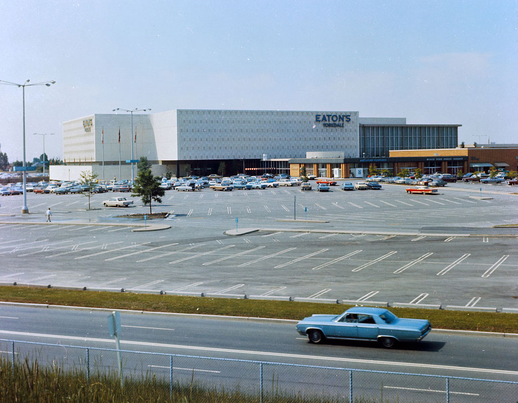 Colour view of large shopping centre with parking lots. Sign saying Eaton's Yorkdale visible on the building. Road in the foreground with blue car.