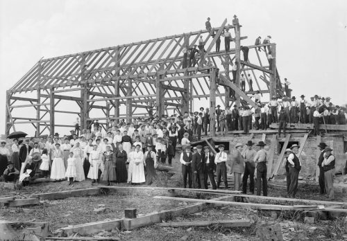 Crowd of people in front of an unfinished timber barn structure.