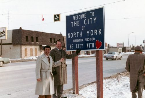 2 men pointing to a street sign which reads Welcome to the City of North York population 563 000 the city with heart